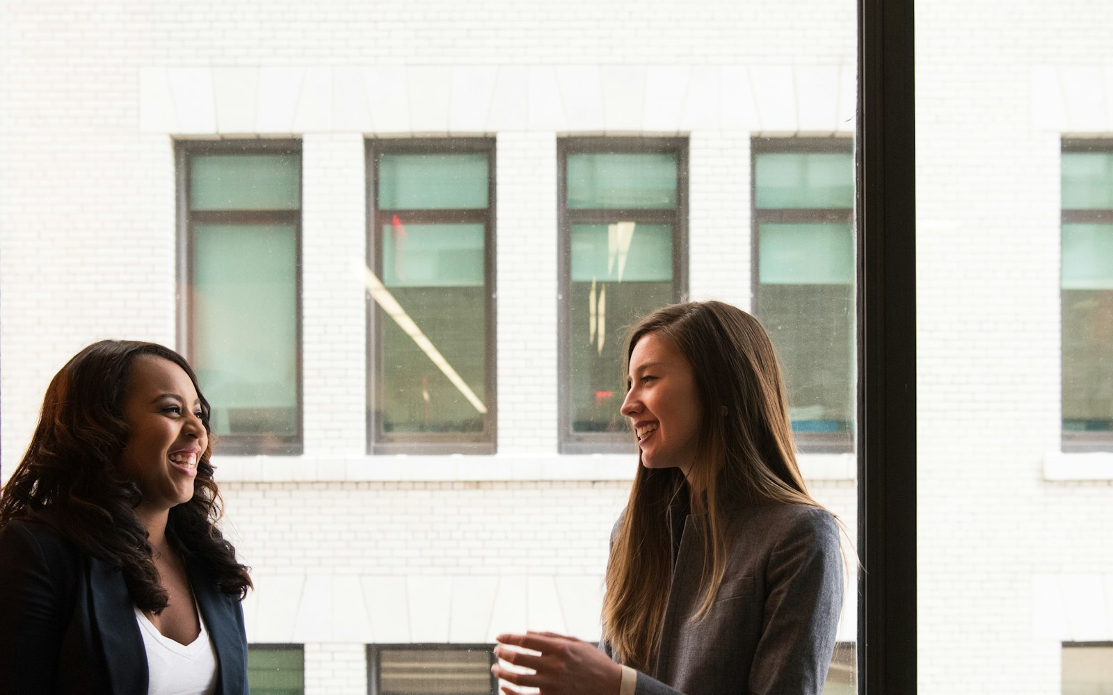 Dos personas conversan sonriendo junto a un ventanal, con luz natural.